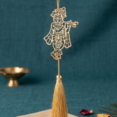 Gold polished brass Krishna flute hanging decor with tassel displayed on teal blue fabric background with peacock feather and brass diya styling.