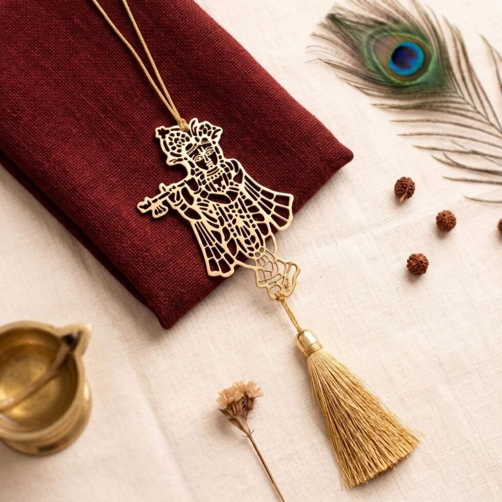 Gold polished Krishna brass hanging decor placed on cream and barn red fabric background with tassel and peacock feather.